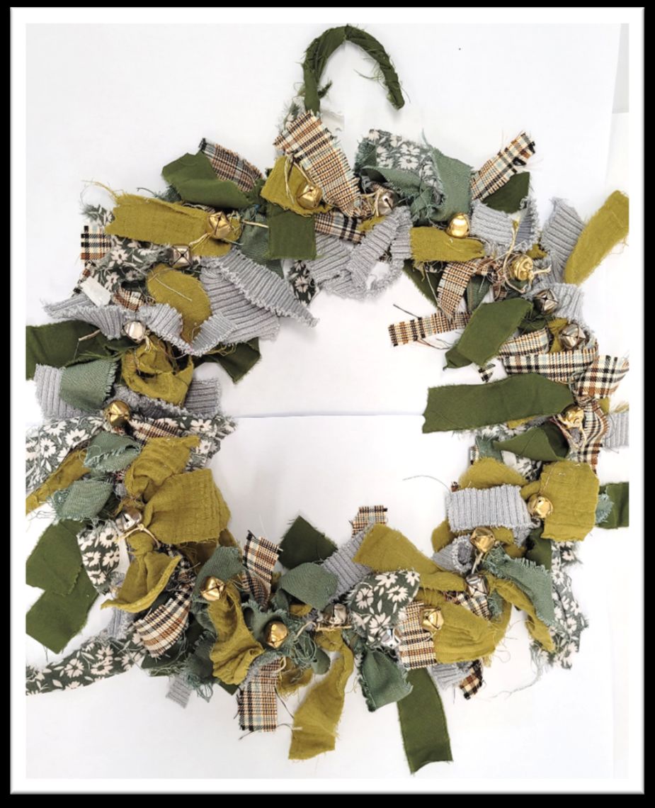 An upcycled circular wreath is crafted by tying numerous strips of earth-toned green, grey, plaid, and floral fabric scraps together, punctuated by small golden jingle bells throughout the frame